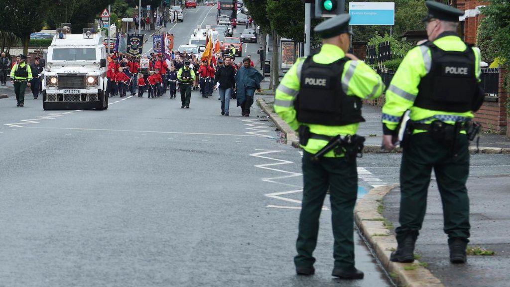 Police in the forground with their back to the camera. In the background a parade of people in red uniform led by a police land rover