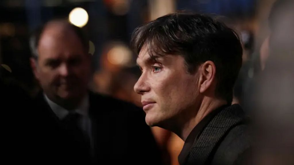 Cillian Murphy is pictured side-on, he is wearing a dark-coloured jacket and a dark-coloured shirt. Only his head and shoulders are seen. There are other people in the background, out of focus.