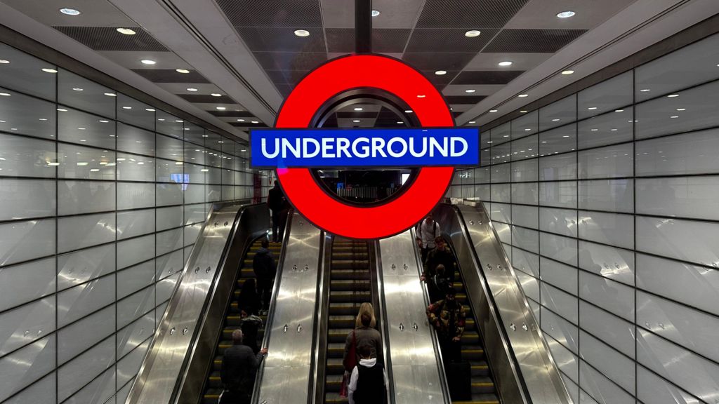 A large Tube roundel hangs above a bank of three escalators as Euston underground station. There are white tiles on the wall and people are visible riding the escalators.