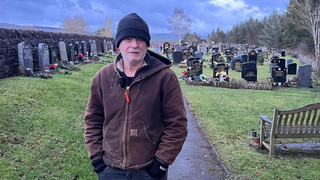 Andy wearing black woollen hat, dark brown top, looking at camera.
Graveyard with grave stones behind him