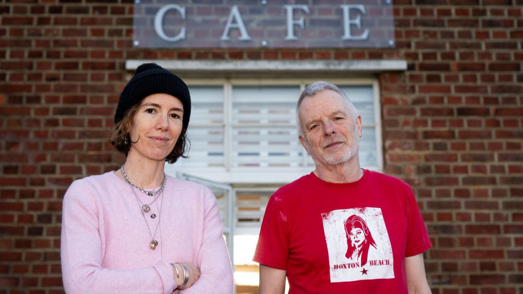Emma Fernandez and Patrick Matthews standing in front of the lido cafe. Emma wears a pink cardigan and a black beanie hat, and Patrick wears a red t-shirt.
