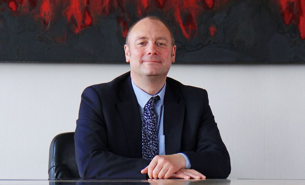 Darren sits at a desk with his hands folded on top of one another. He smiles at the camera and is wearing a dark blue suit with a floral navy tie.