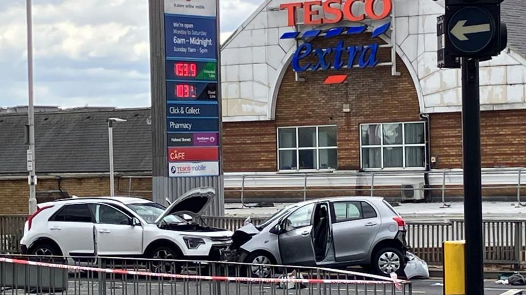 A Tesco Extra can be seen in the background of the shot along with the two collided vehicles before a metal fence. A police cordon is closing off the area.