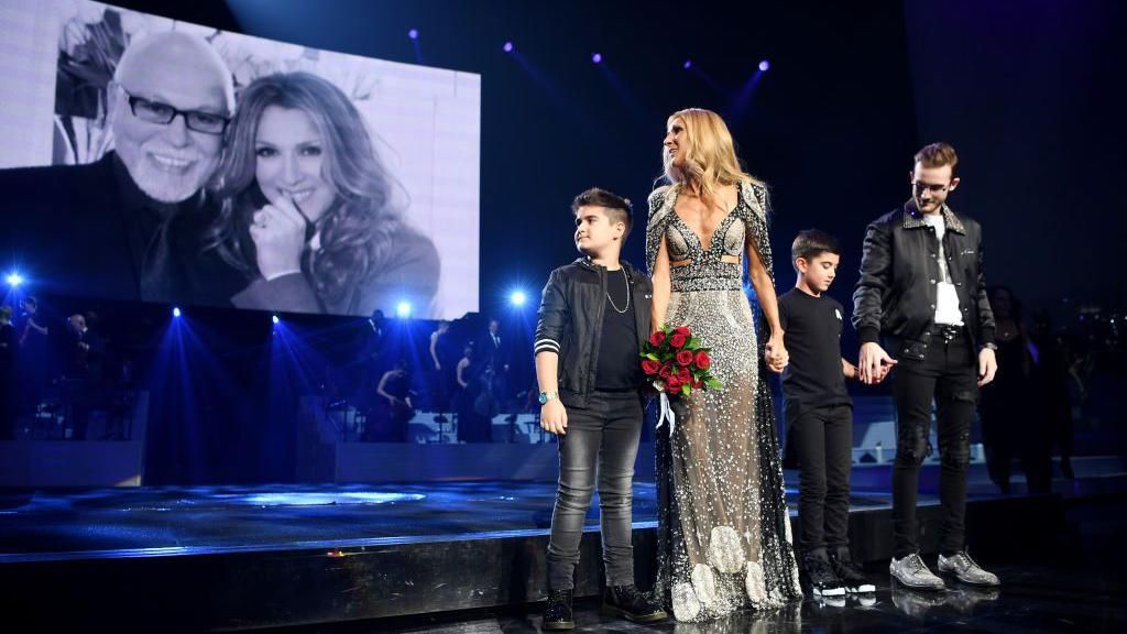 Celine Dion holding hands with her children on stage, with a photo of her and late husband René Angélil projected on the screen