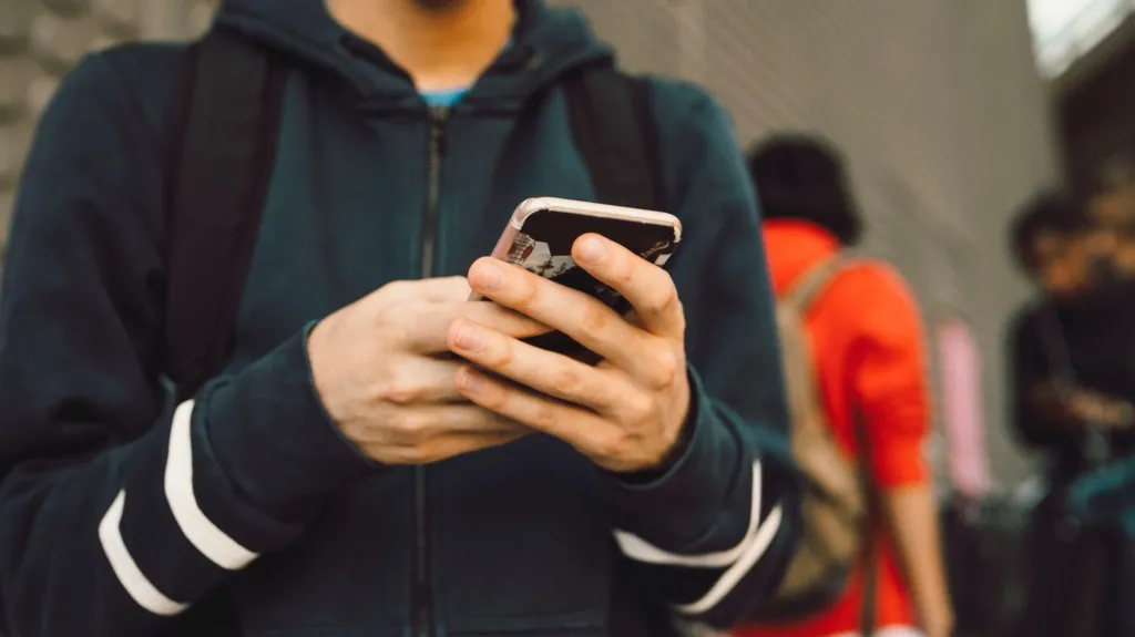 Un joven sostiene un teléfono móvil con ambas manos, vistiendo una sudadera con capucha. La imagen está tomada en un entorno urbano, con otras personas desenfocadas en el fondo.