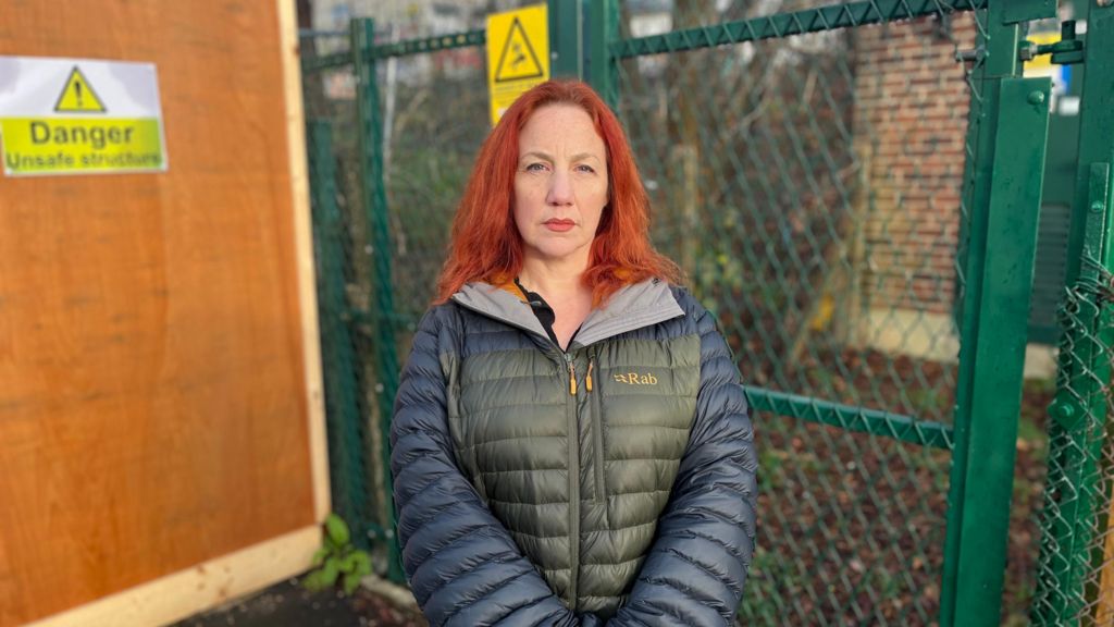 A woman with red hair wearing a warm green and blue coat. In the background is green fencing separating her from the woodland and a building with a "Danger: Unsafe structure sign".