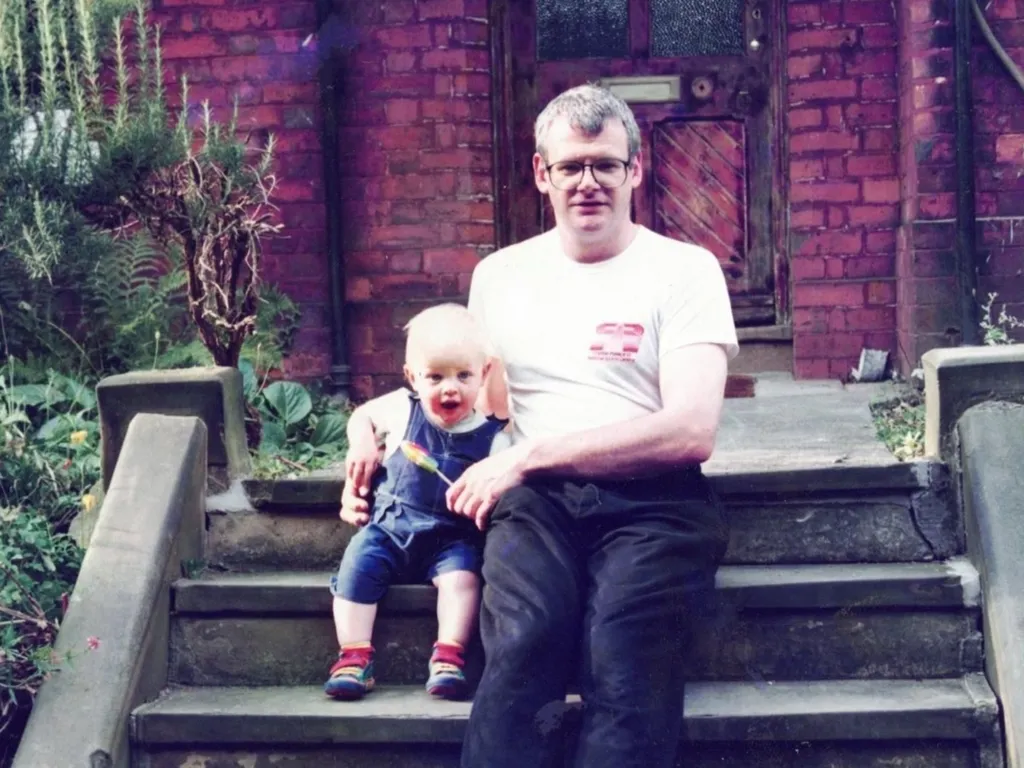 Andrew, in dark trousers and a white T-shirt, sits on garden steps with his toddler son Harry, who is wearing blue dungarees.