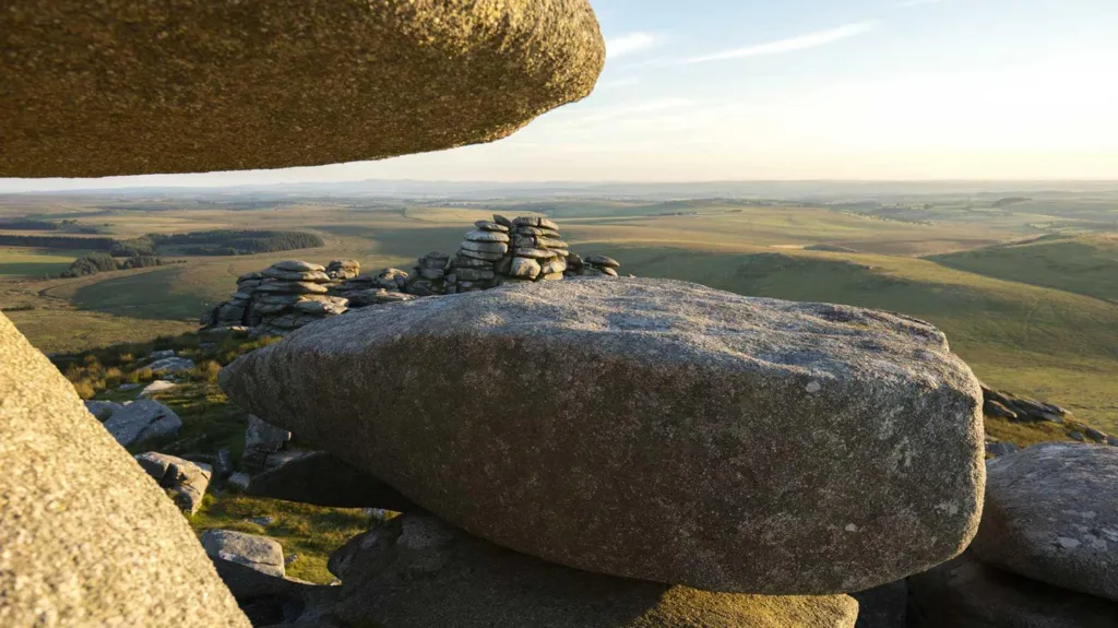 Rough Tor on Bodmin Moor on a clear day. There are giant granite boulders balanced on top of each other and beyond are the grassy moors which stretch in to the distance.