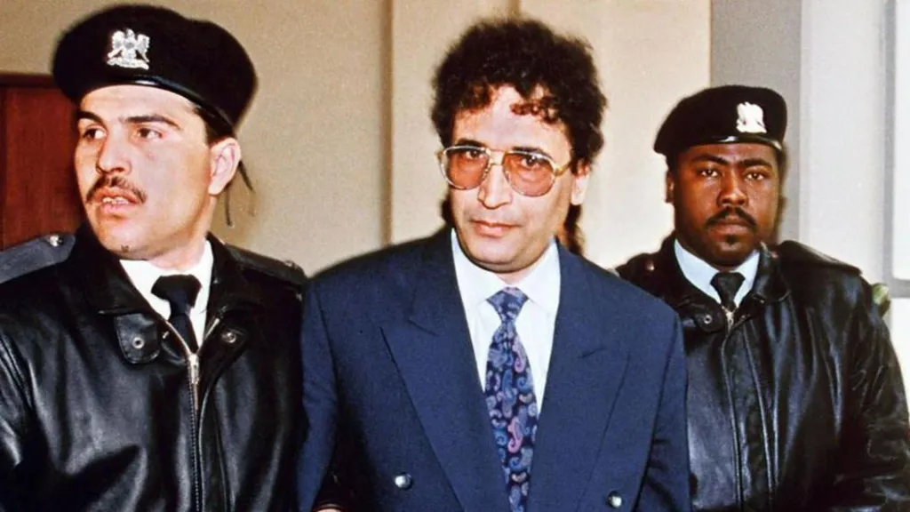 Abdelbaset Ali Mohamed al-Megrahi, wearing a blue suit over a white shirt and a floral tie, with two security officers in black leather jackets and berets.