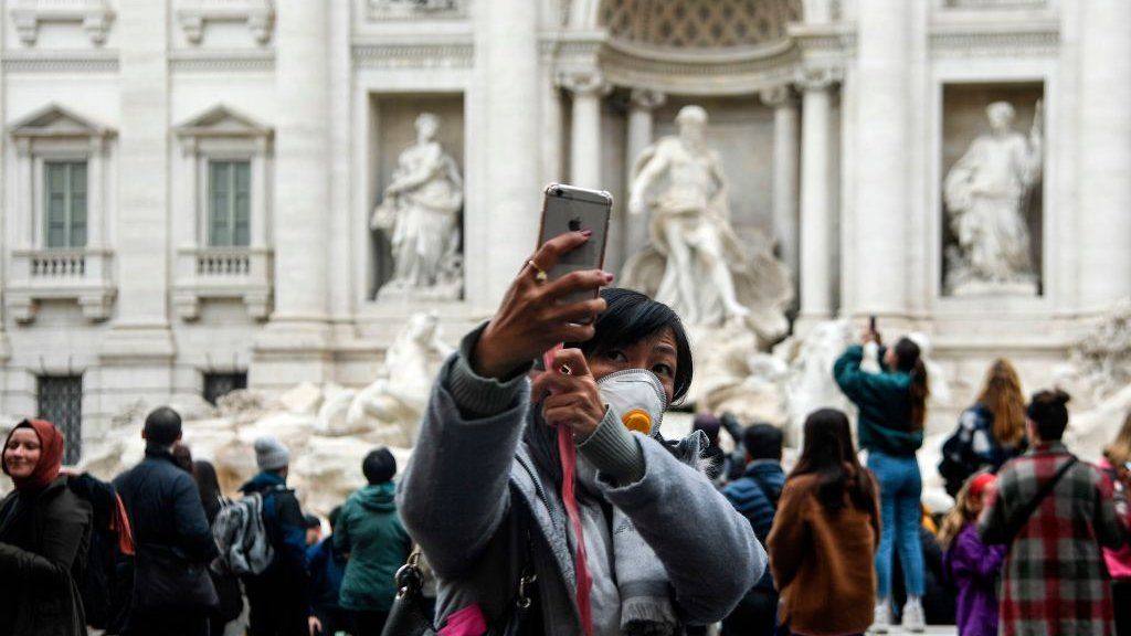 A Chinese tourist by the Trevi fountain in Rome, taking a selfie while wearing a face mask
