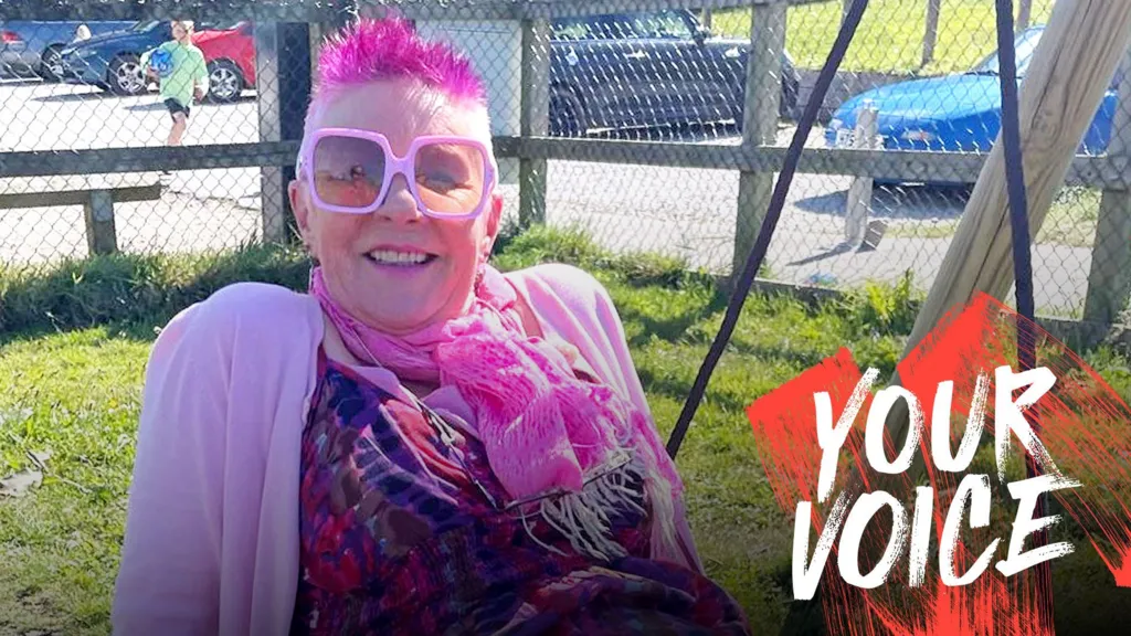 Annie Haslam, with short pink spiky hair, pink glasses, pink scarf and pink cardigan, sits on a big swing outside