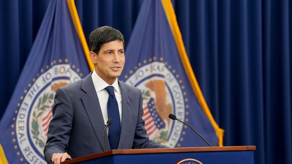 Kevin Warsh stands at a podium giving a press conference, wearing a navy suit, navy tie and white shirt, flanked by flags with the crest of the Federal Reserve.