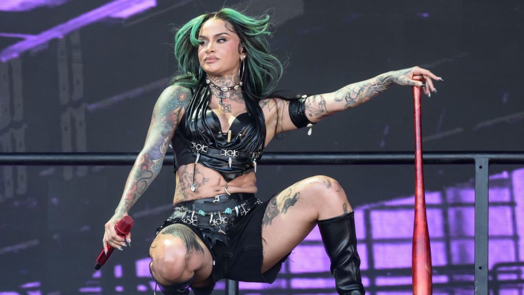 Kehlani performs on stage wearing a black leather bra top and shorts, paired with black knee-high boots. She has black hair with green highlights. She is crouched down on the stage, leaning on a red baseball bat.