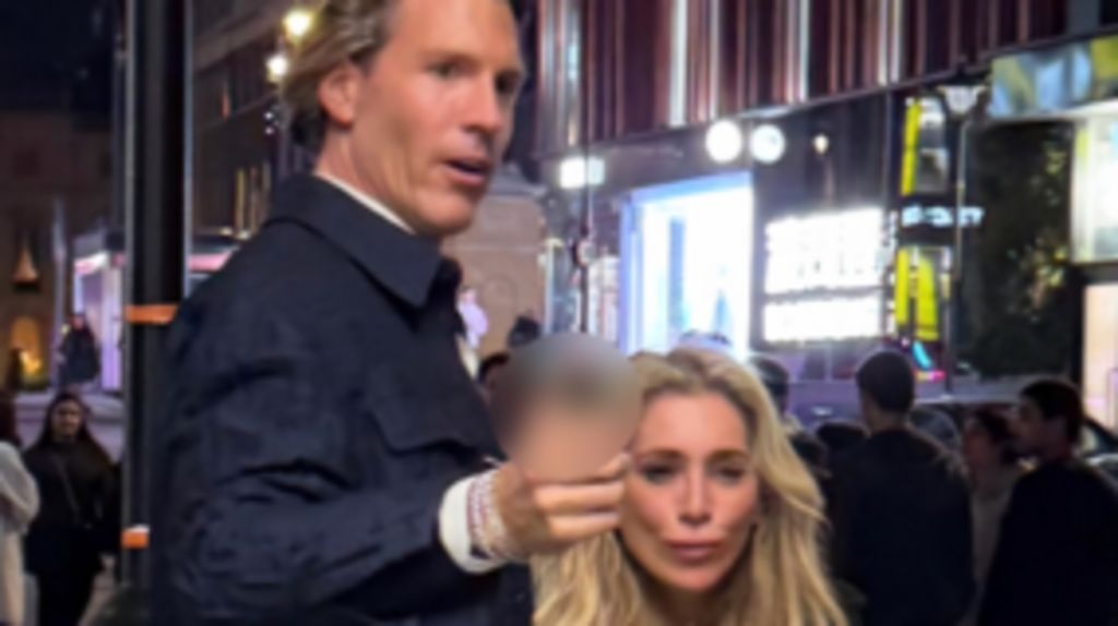 A CCTV image showing a tall man with brown hair wearing wearing a smart dark blue jacket, gesturing forward. He is with a woman with long, blonde hair 