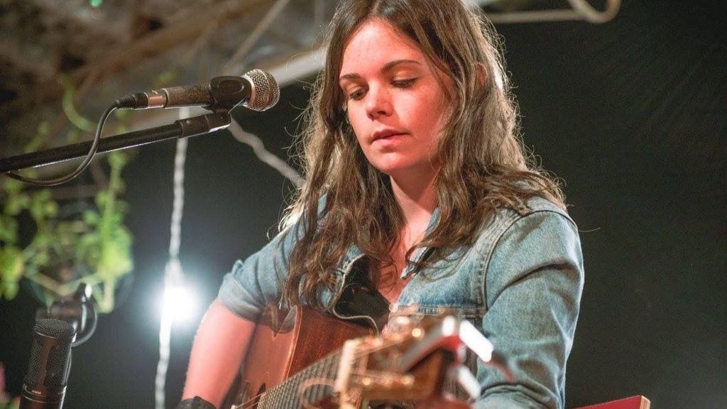 Jessie Reid sitting in front of a microphone with a guitar. She has long, brown hair and is wearing a blue denim jacket