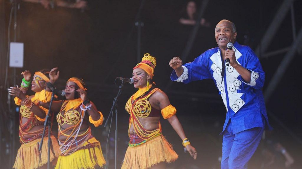 Femi Kuti dancing in a blue suit with three dancers next to him wearing orange and yellow outfits