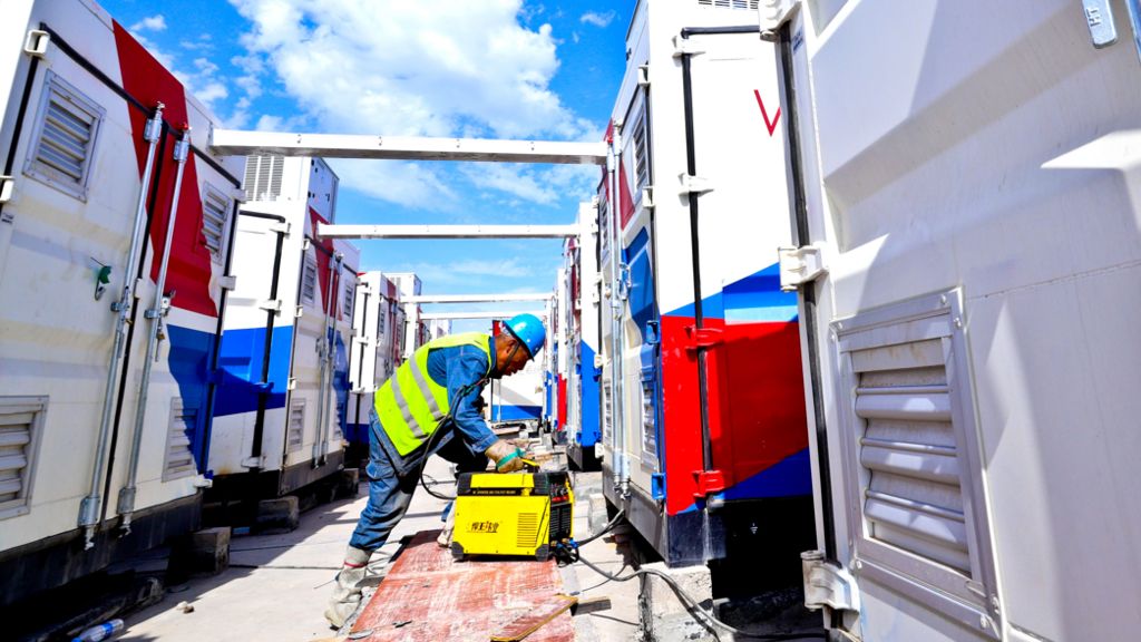 A man in a hi-viz jacket with blue overalls and a blue hard hat working at a battery energy storage system, he is surrounded by large white metal structures