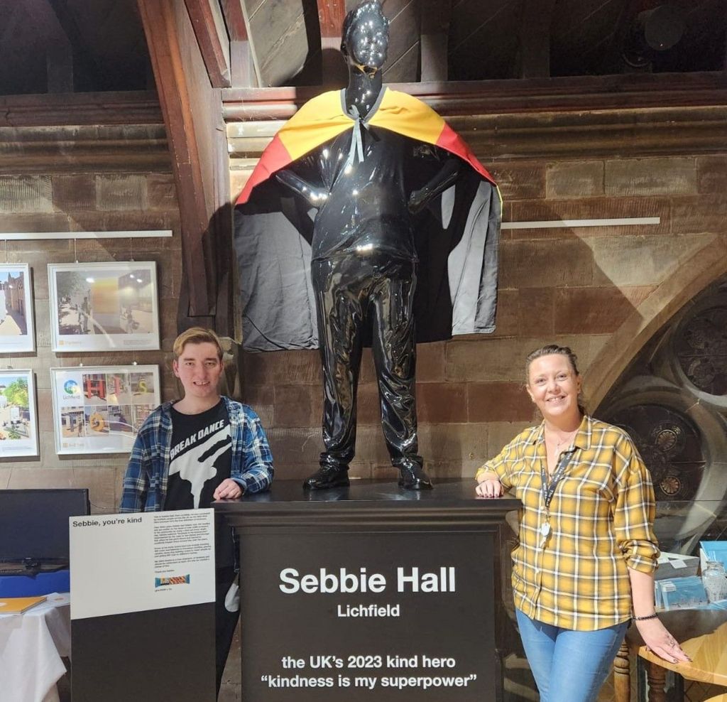 A man in a black tshirt with a blue checked shirt over the top of it stands next to a statue with a woman on the other side of the statue wearing a yellow checked shirt