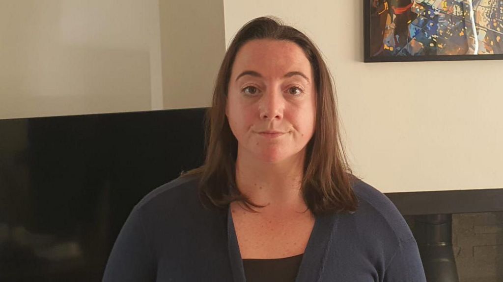 Beth Rodgers, a 32-year-old woman. She is standing in her living room wearing a navy cardigan and black top.