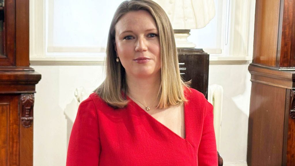 Barrister Charlotte Proudman is seated and looks into the camera with a neutral expression. She has straight, shoulder‑length fair hair and is dressed in bright red. There is polished wooden furniture either side of her and a white sculpted bust behind. A window casts soft natural light.