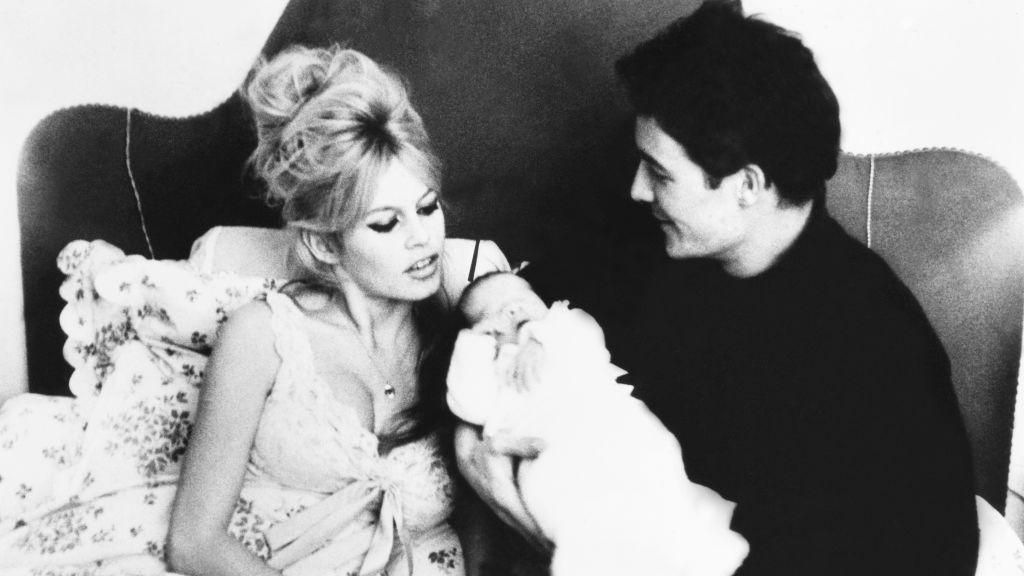 Brigitte Bardot sits in bed, wearing a light-coloured nightie, her blonde hair half piled on her head, wrapped in floral bedsheets, while Jacques Charrier, wearing a dark shirt and trousers, holds their newborn baby Nicolas in a white outfit.