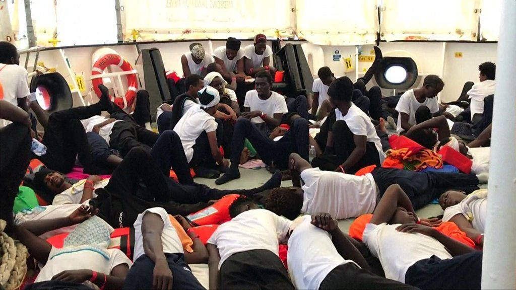 Dozens of migrants in identical white t-shirts and dark trousers lie on the deck of the Aquarius
