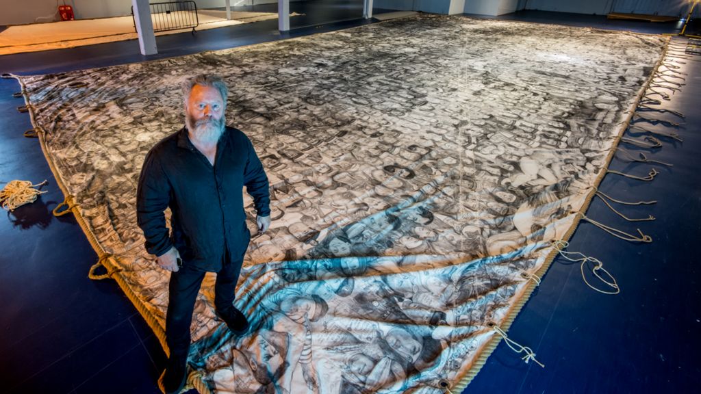 A man with a beard stands in front of a large boat sail. The sail is laid out on the floor and is covered in charcoal drawings of people's faces