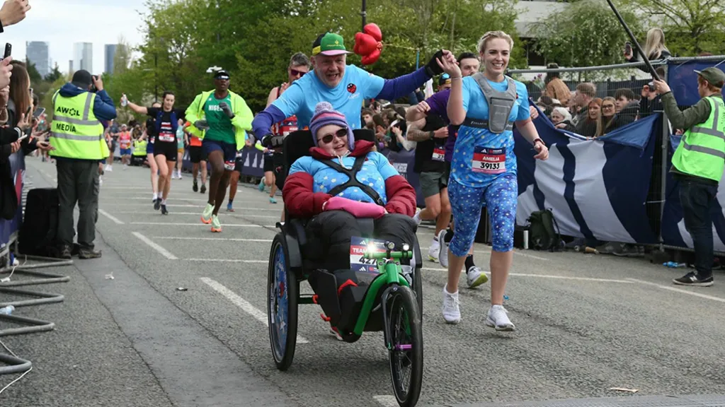 Stephan is pushing Chloe in her race wheelchair during a race. He is wearing a blue t-shirt and green and black cap. A woman is holding Stephan's hand while she runs alongside him. There is a crowd lining the route on both sides, as wel as other runners in the background