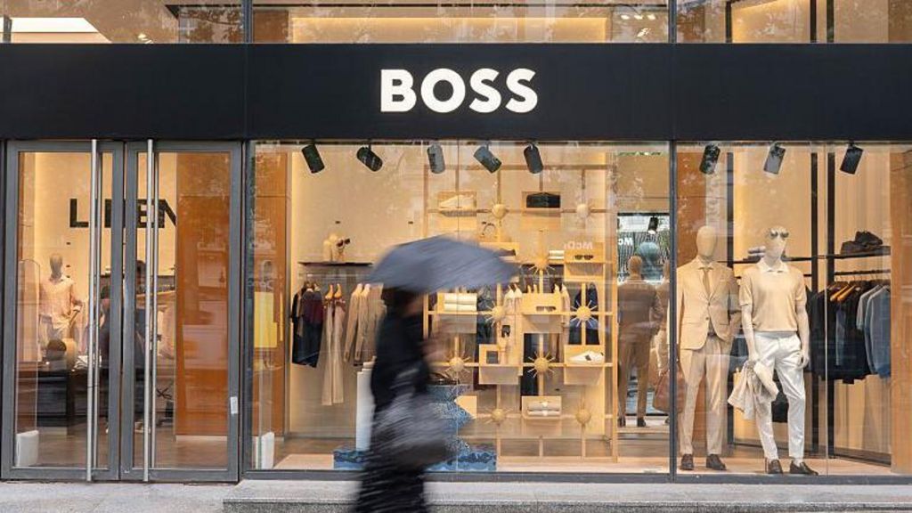 The entrance of the Hugo Boss flagship fashion store in Paris, France. In the foreground, a blurred woman is walking past with an umbrella. Inside the shop, mannequins, shirts, shoes, and trousers are visible.