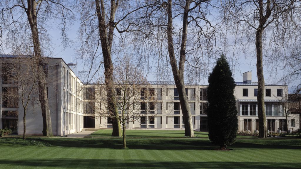 long, grey college building with elderly trees in the foreground and a well tended lawn