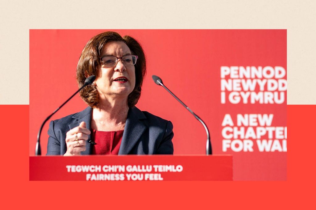 First Minister of Wales Baroness Eluned Morgan addresses an audience of members and candidates at Newport Market. She is wearing a dark suit over a red top and stands in front of a red backdrop that reads "PENNOD NEWYDD I GYMRU / A NEW CHAPTER FOR WALES." In front of her is a lectern with two microphones. On the front of the lectern is written: "TEGWCH CHI'N TEIMLO / FAIRNESS YOU FEEL."