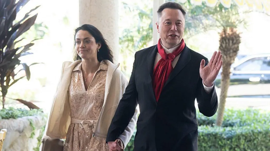 Shivon Zillis, wearing a pink metallic dress and cream jacket, walks hand in hand with Musk, who wears a red scarf, white shirt and black suit, as he waves to photographers.