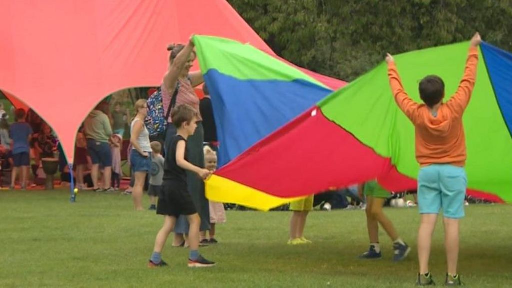 A group of children and an adult can be see playing on grass with a large multi-coloured parachute. Behind them is a large red marquee with a number of children and adults inside. 