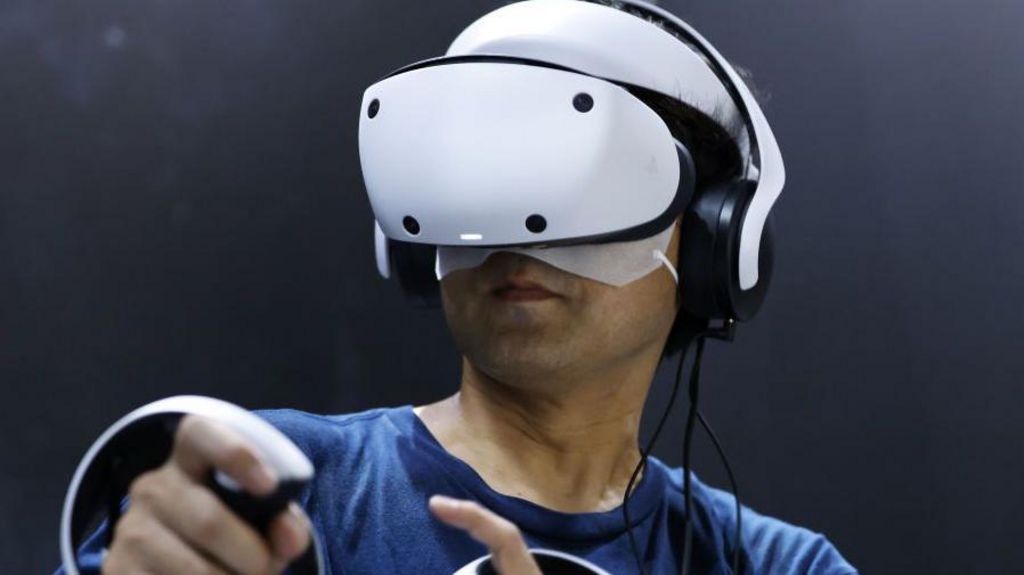 A man uses the PlayStation VR 2. It is larger than the other two headsets, and it is a sleek white curved shape.