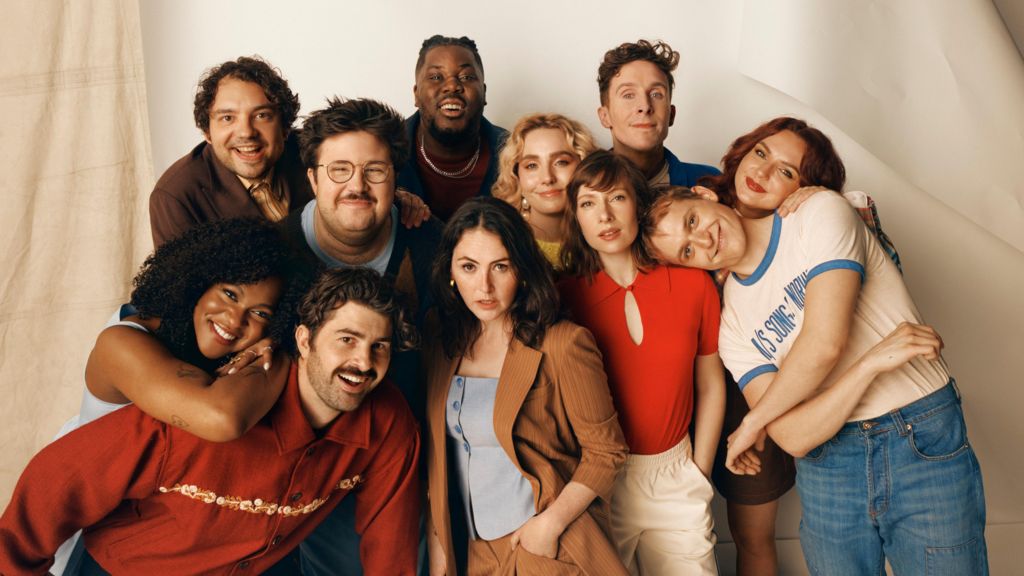 Posed picture of SNL UK cast smiling for the camera. They are standing against a beige backdrop.