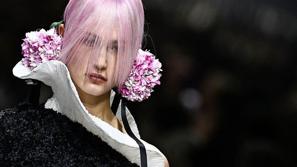 A model with light pink hair and a long fine fringe covering her face wears a black dress with a white wavy collar - purple and white floral balls are at her ears