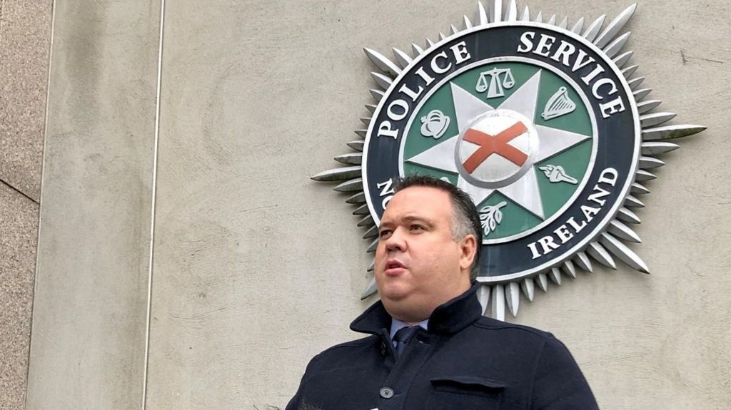 A man with dark hair in front of a Police Service of Northern Ireland logo