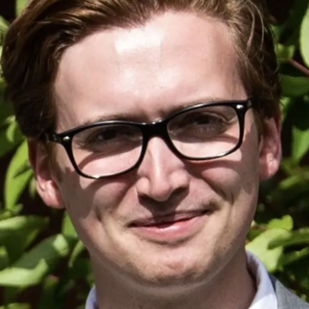 Fletcher Mumford, a young man with short, auburn hair and black-rimmed glasses looks towards the camera