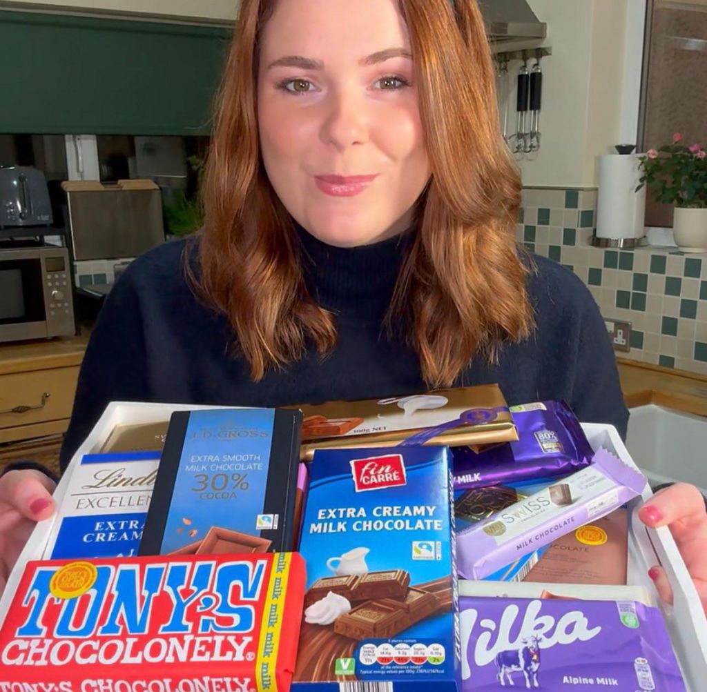 Becca Amy Stock is smiling and looking at the camera. In front of her she is holding a tray of milk chocolate bars, all different brands.