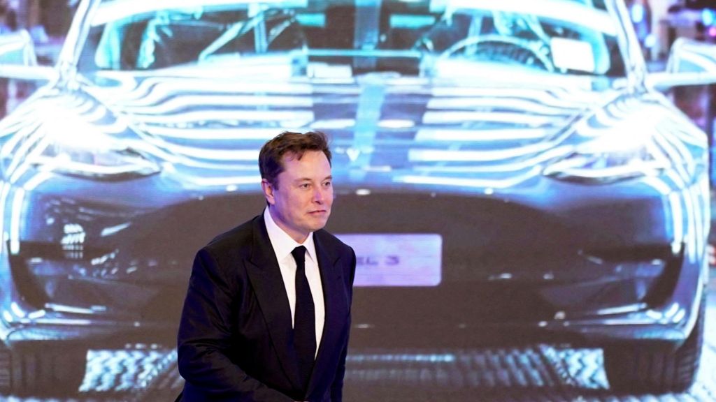 Elon Musk, wearing a black suit, white shirt and black tie, walks in front of a large display showing a Tesla Model 3 at an event in 2020 