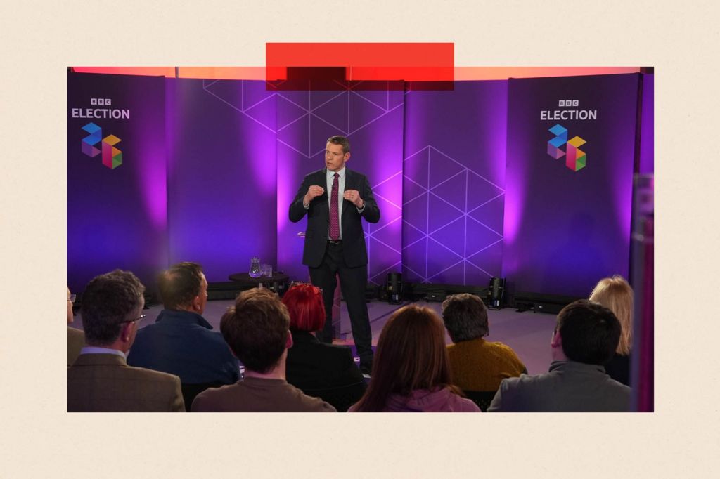 Plaid Cymru leader Rhun ap Iorwerth at a BBC Wales Ask the Leaders debate in Haverfordwest, Pembrokeshire. He is wearing a dark suit and a white shirt with a purple tie and stands in front of an audience, whose heads are visible from the rear. The words "BBC ELECTION 26" appear in two places on a screen behind him.