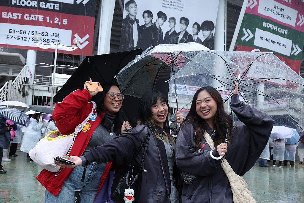 Three BTS fans smile and pose for the camera outside the Goyang Stadium ahead of the first date of the band's world tour. They are shielded under umbrellas, while other concertgoers in the background are covered in plastic raincoats. 