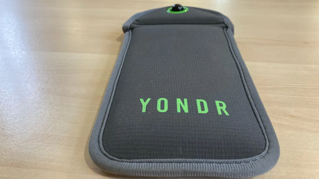 A grey pocket sleeve wallet sits on a wooden table. On it is the company's name Yondr in green colouring. It is designed to store a mobile phone device with a magnetic seal which can be reopened at a later time or date