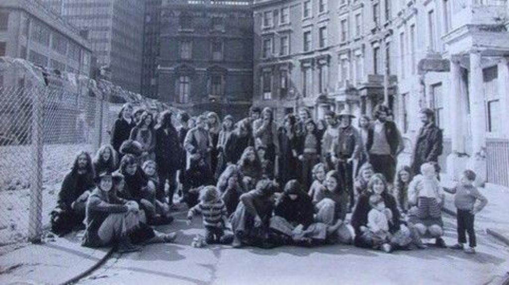 A black and white photo from the 1980s of a group of adults and children sitting on the road and pavement in front of a row of houses