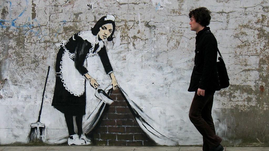 Sweep It Under The Carpet on a wall in Camden, depicting a maid in black and white with a dustpan and brush appearing to pull up a painted wall to expose the bricks behind