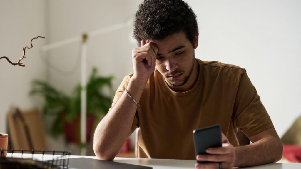 A young man looks at his phone while sitting at a computer in his home. He looks weary.