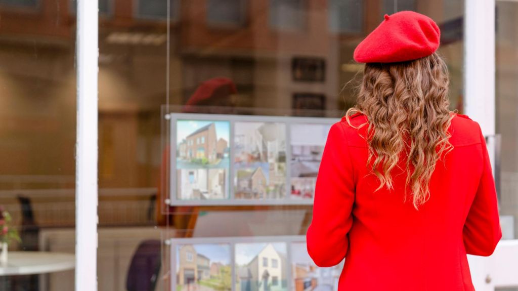 Woman wearing bright red beret and red coat faces an estate agent's window advertising homes for sale 