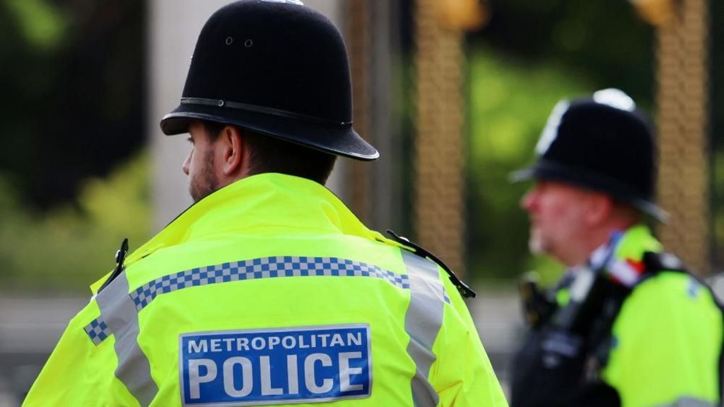 Two police officers in high vis face away from the camera and they have "Metropolitan Police" printed on their backs. One man has his back to the camera and another looks to the left. Both wear helmets.