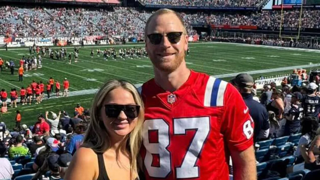 Seamus Culleton and his wife standing at an American football game. He has a red shirt on with the number 87 in white. They are both wearing black sunglasses. The pitch be hind them is green. There is a crowd in the stands. 