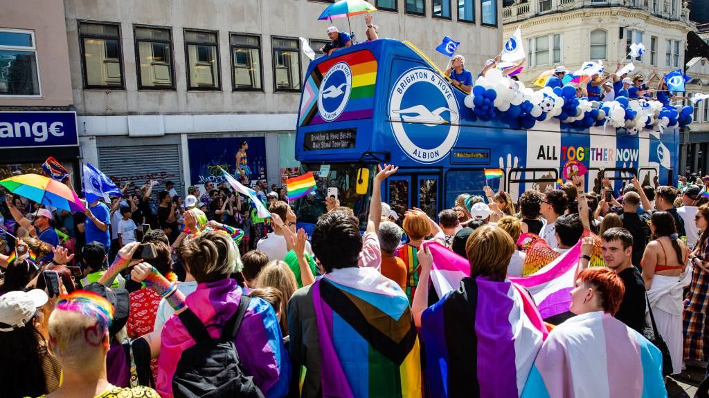 A bus driving through the streets of Brighton with crowds waving rainbow flags